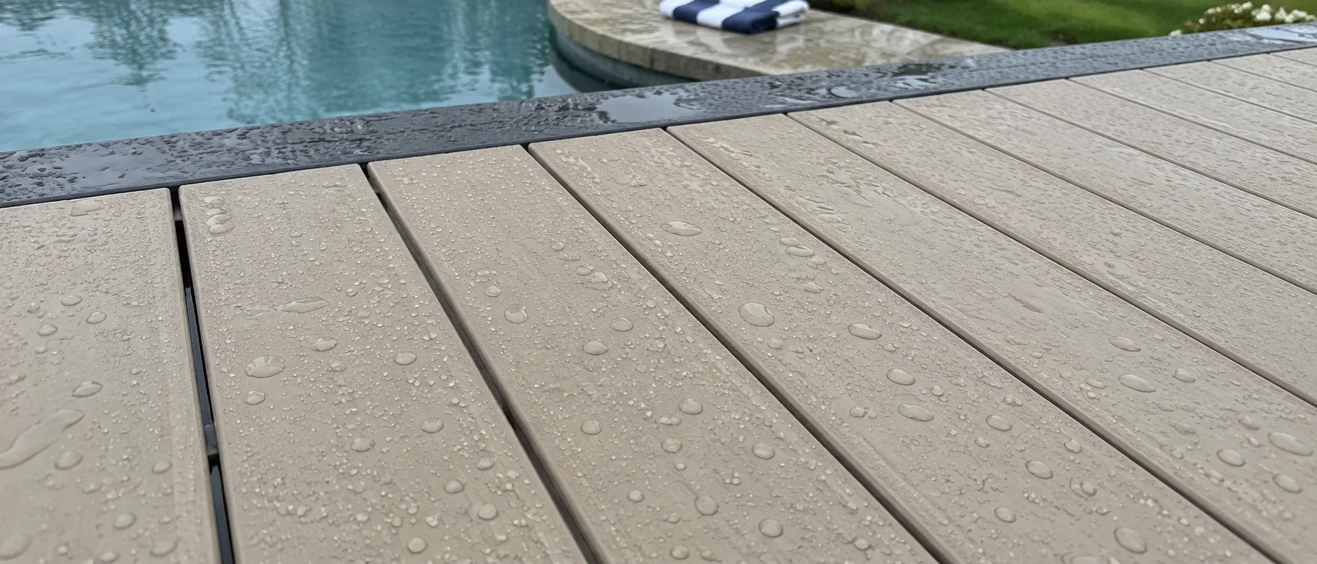 Rain beading on the surface of pale capped PVC decking rather than soaking in, with the edge of a swimming pool beyond