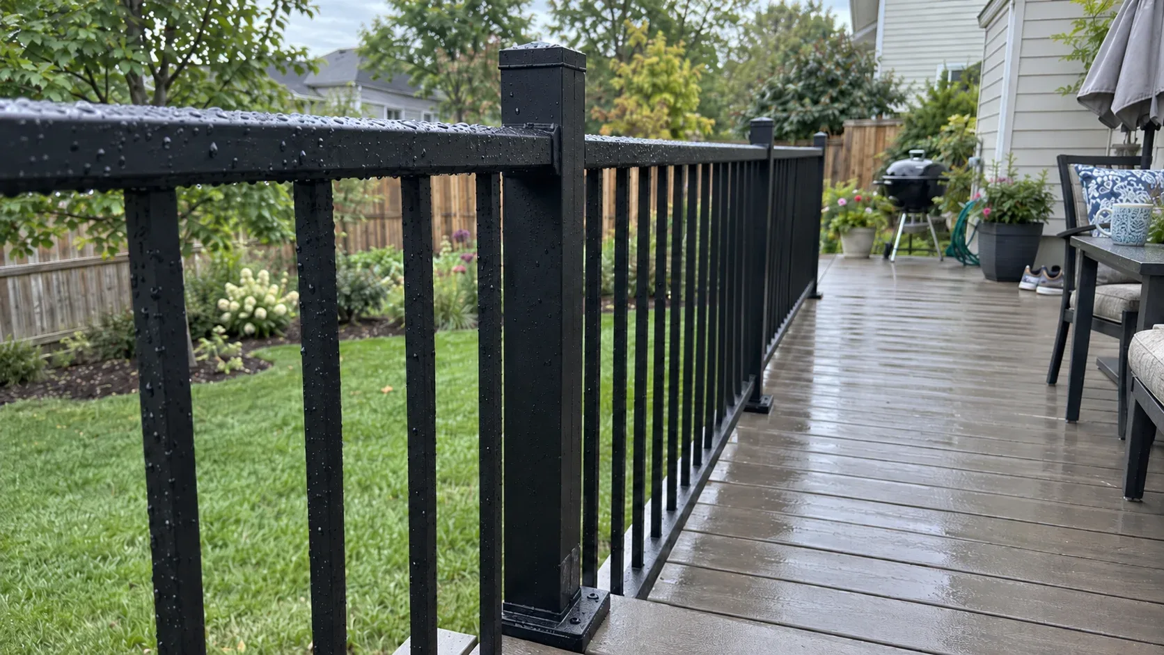 A matte black aluminium deck railing after rain, with evenly spaced square balusters and the post base plate seated on the decking